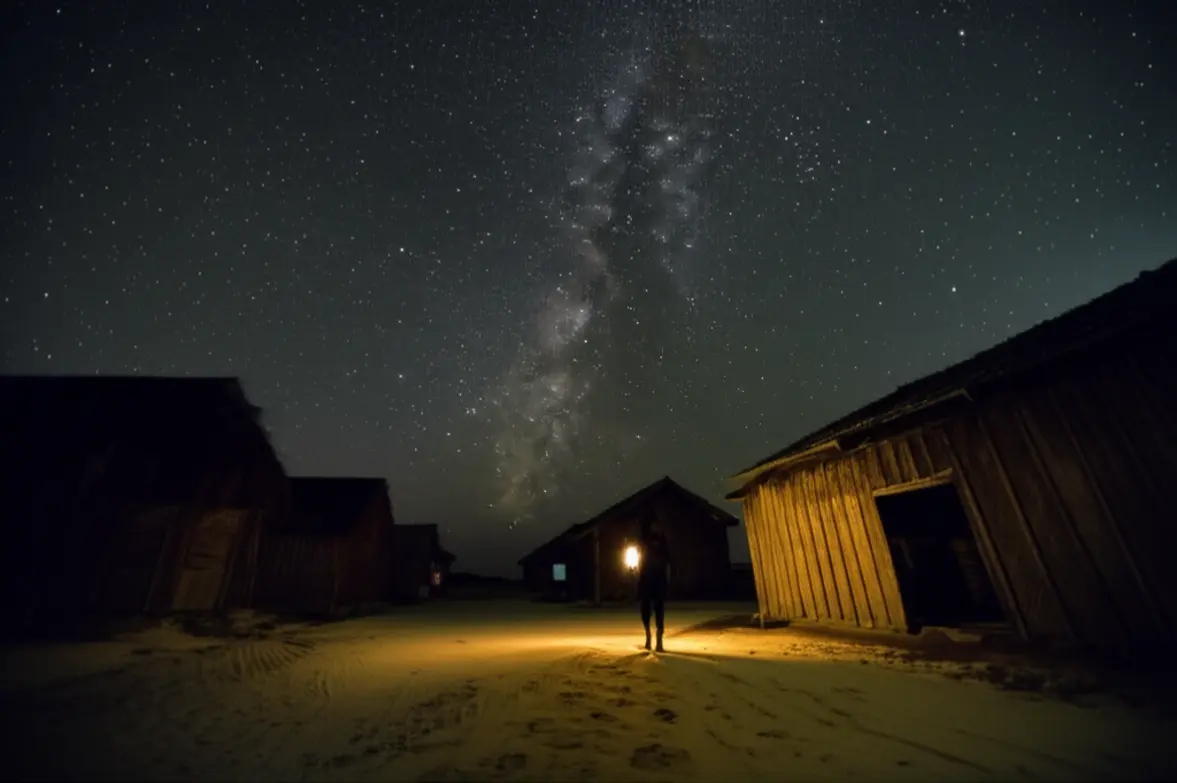 Céu noturno com Via Láctea e uma pessoa caminhando com lanterna na rua de areia em Atins.