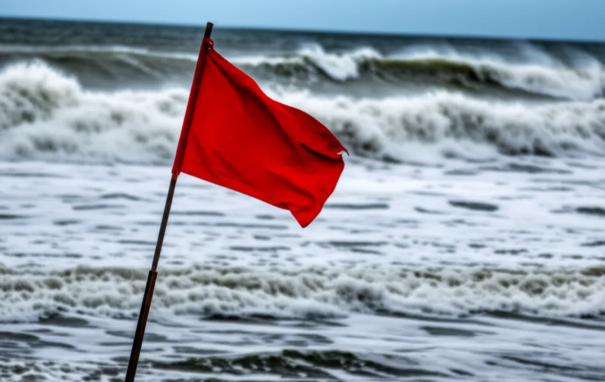 Bandeira vermelha de perigo na areia com mar revolto ao fundo em Saquarema.