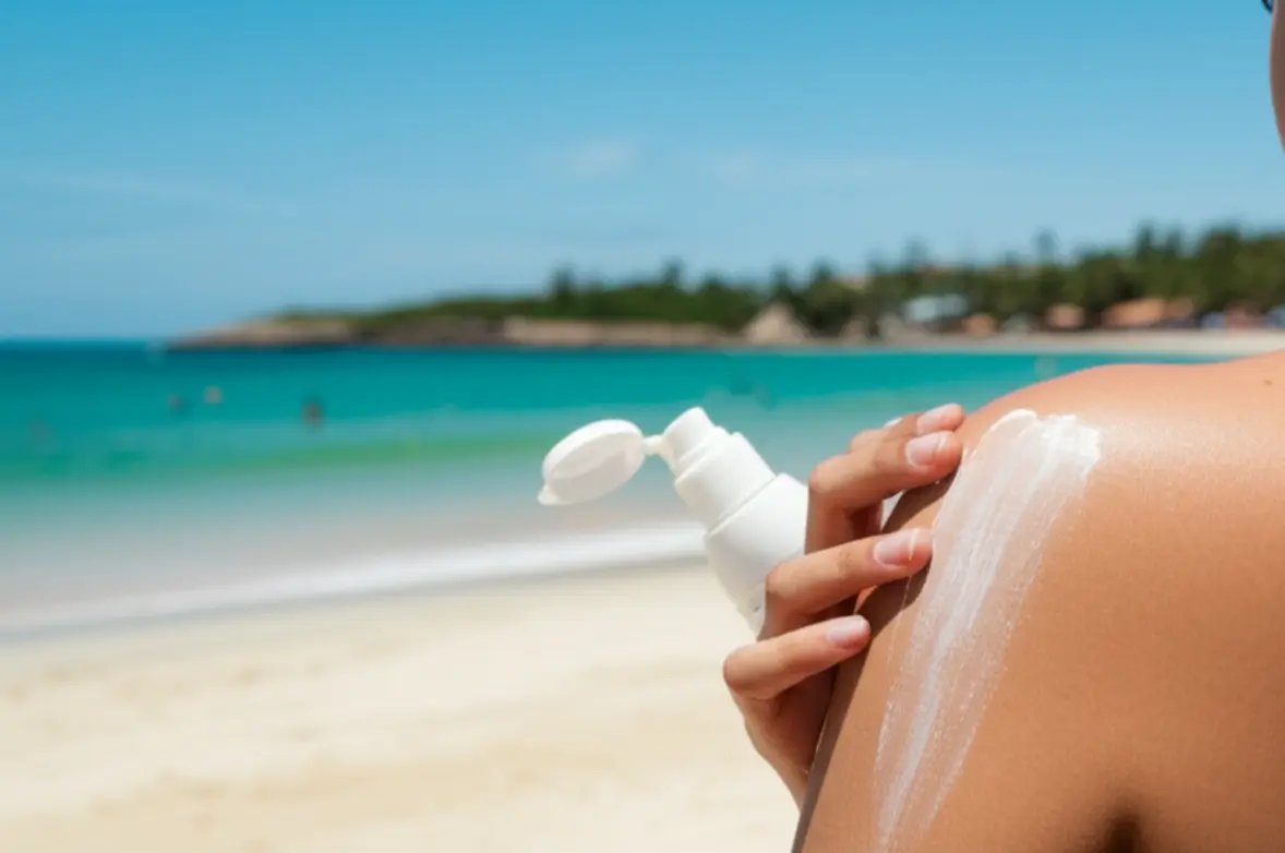 Pessoa aplicando protetor solar na praia, com água turquesa ao fundo, sob sol forte.
