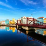 Fotografia de Recife com edifícios coloridos e pontes sobre o rio, sob o sol.