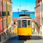 O icônico bonde amarelo elétrico 28 subindo uma ladeira em Lisboa com o Rio Tejo ao fundo.