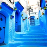 Rua estreita e icônica de Chefchaouen com paredes azuis e vasos de flores coloridos.